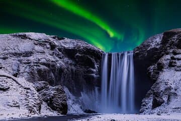 Skogafoss Waterfall flows beneath vibrant green northern lights lighting up the night sky.