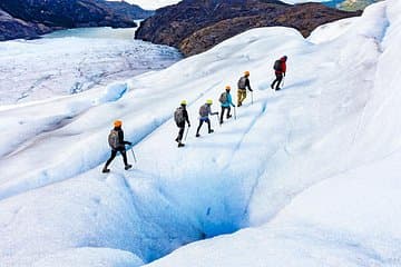 A small group of hikers walks across a vast glacier in Iceland, wearing crampons and carrying ice axes.